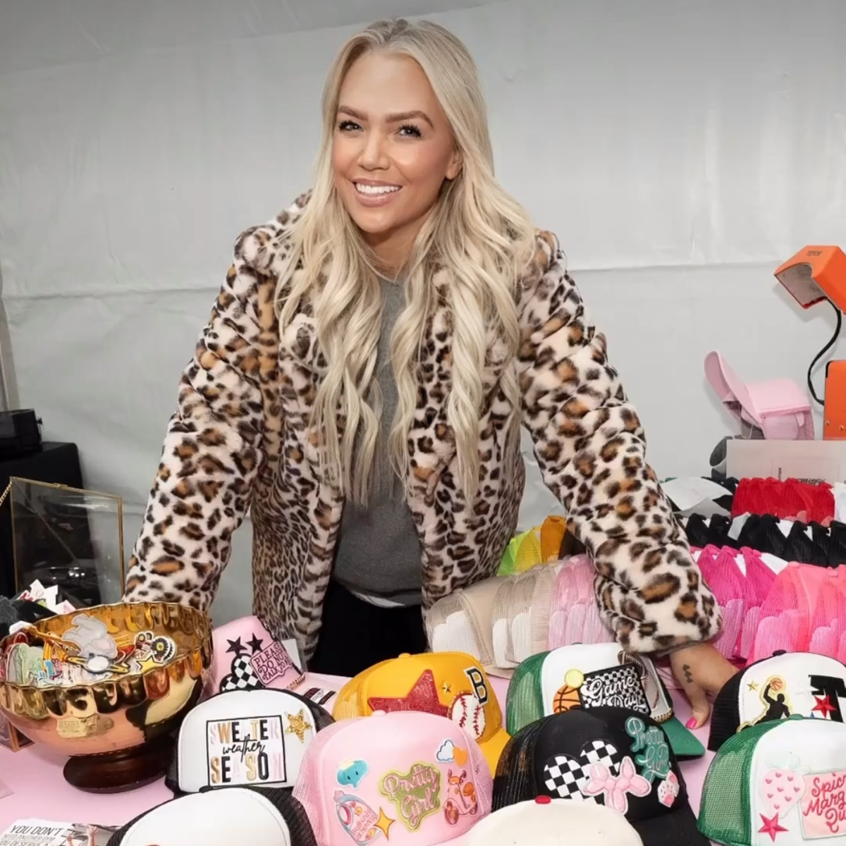 Woman in leopard print jacket standing behind a display of colorful hats and accessories.