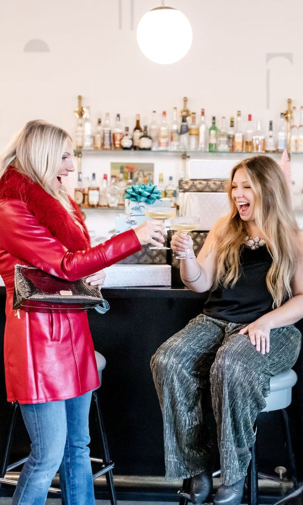 Two women in a bar setting, one in a red jacket and blue jeans, the other in a black top and patterned pants, enjoying drinks. Vixen Collection Seattle, WA
