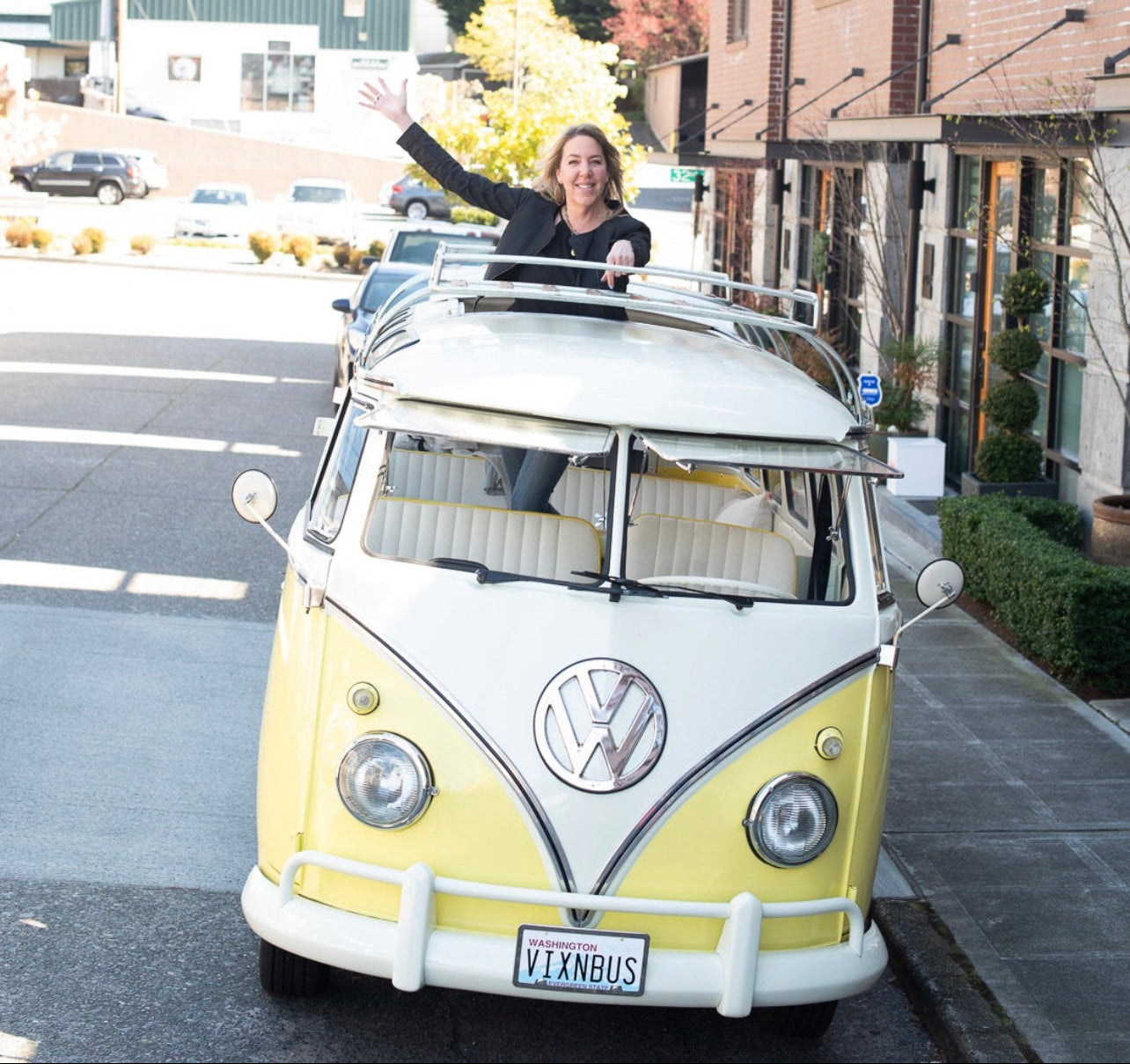 A yellow and white classic Volkswagen Type I bus parked on a city street, with a person standing on the roof and waving. The photo is shown within a mobile app interface with a white border.